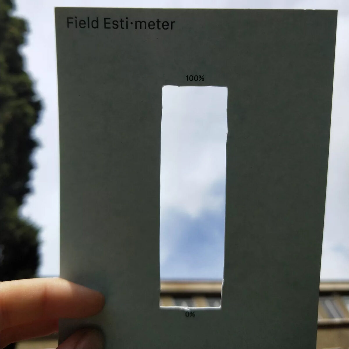 Finger halten eine Postkarte mit der Bezeichnung 'Field-Estimeter'. Die Karte hat ein längliches Loch, markiert mit '100%' oben und '0%' unten. Durch das Loch ist der Himmel und am unteren Rand ein grüner Horizont zu erkennen, was auf eine niedrige Schätzung hinweist.