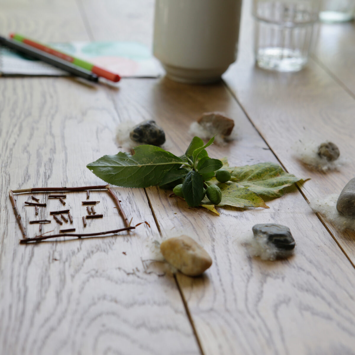 Ein Holztisch mit einer Anordnung aus Stöckchen, die ein geometrisches Muster bilden, umgeben von grünen Blättern, Pollen und einigen Steinen. Im Hintergrund liegen ein Notizbuch, farbige Stifte, ein Becher und ein Wasserglas.